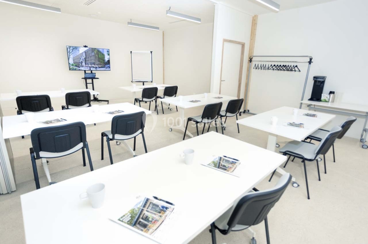Salle de formation lumineuse avec tables blanches, chaises noires, écran, paperboard et porte-manteaux.
