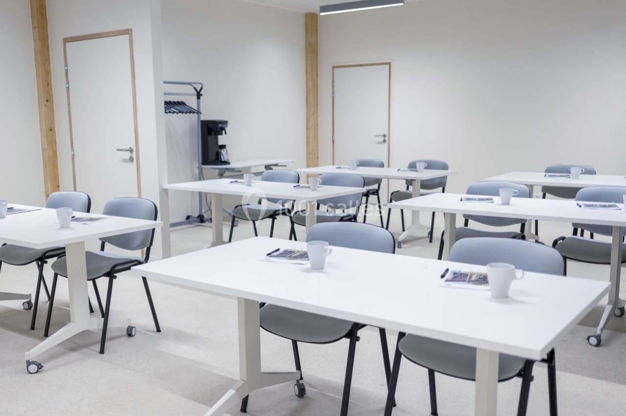 Salle de formation lumineuse avec des tables blanches, des chaises grises et des documents posés sur les tables.