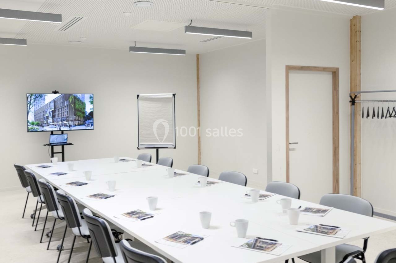 Salle de réunion lumineuse avec table en U, chaises, écran affichant un bâtiment, paperboard et porte-manteaux.