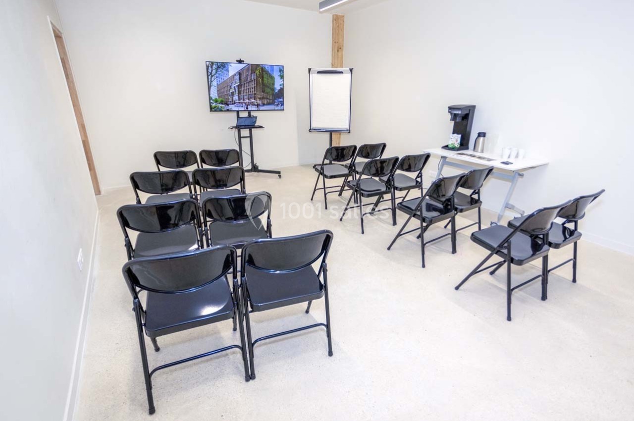 Salle de réunion avec chaises noires alignées, écran, paperboard et table équipée d'une machine à café et gobelets.