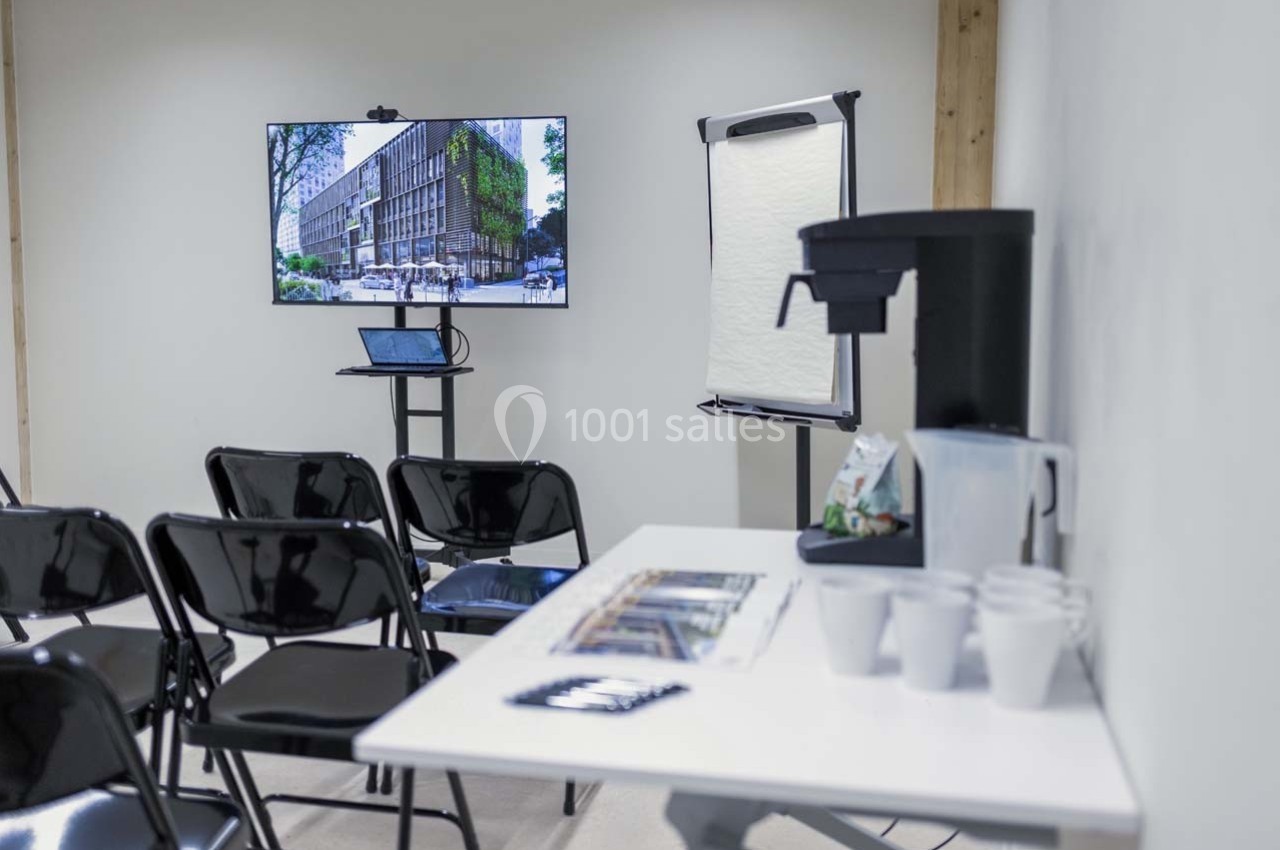 Salle de réunion avec chaises, écran affichant un bâtiment, table avec cafetière, gobelets et bloc-notes.