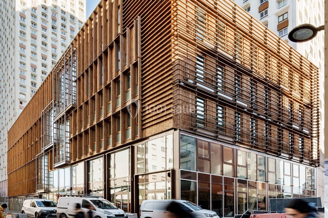Façade moderne d'un bâtiment en bois et verre entouré d'immeubles, avec des passants et des véhicules en mouvement.