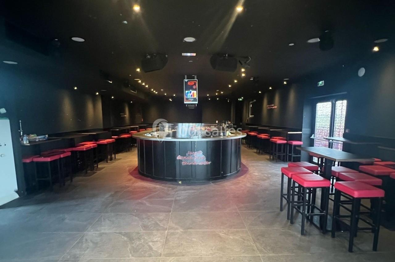 Salle de bar sombre avec un comptoir central circulaire, tabourets rouges et éclairage tamisé.