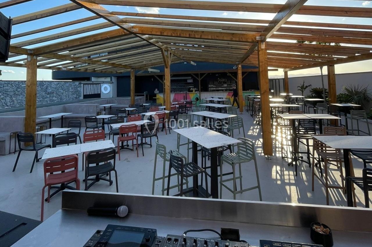 Terrasse extérieure avec tables et chaises sous une pergola en bois, vue depuis une table de mixage au premier plan.