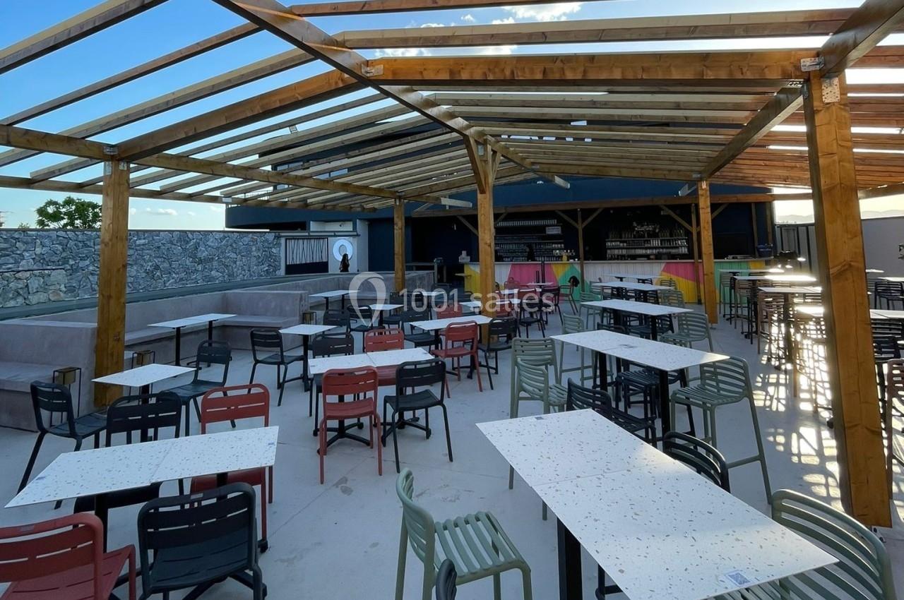 Terrasse extérieure avec tables et chaises colorées sous une structure en bois, bar en arrière-plan.