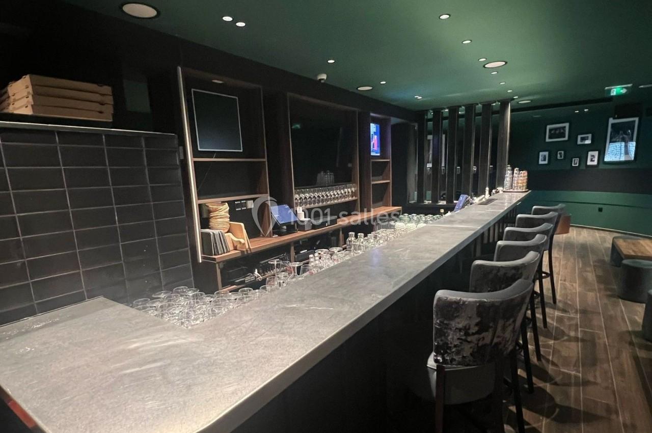 Bar moderne avec comptoir en pierre, tabourets, étagères garnies de verres et éclairage tamisé.