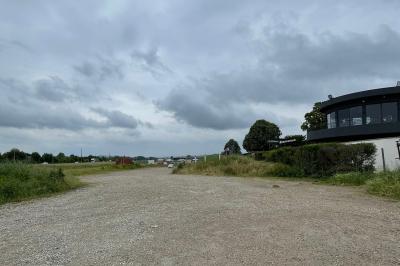 Chemin de gravier bordé de végétation, menant à un bâtiment moderne sous un ciel nuageux.