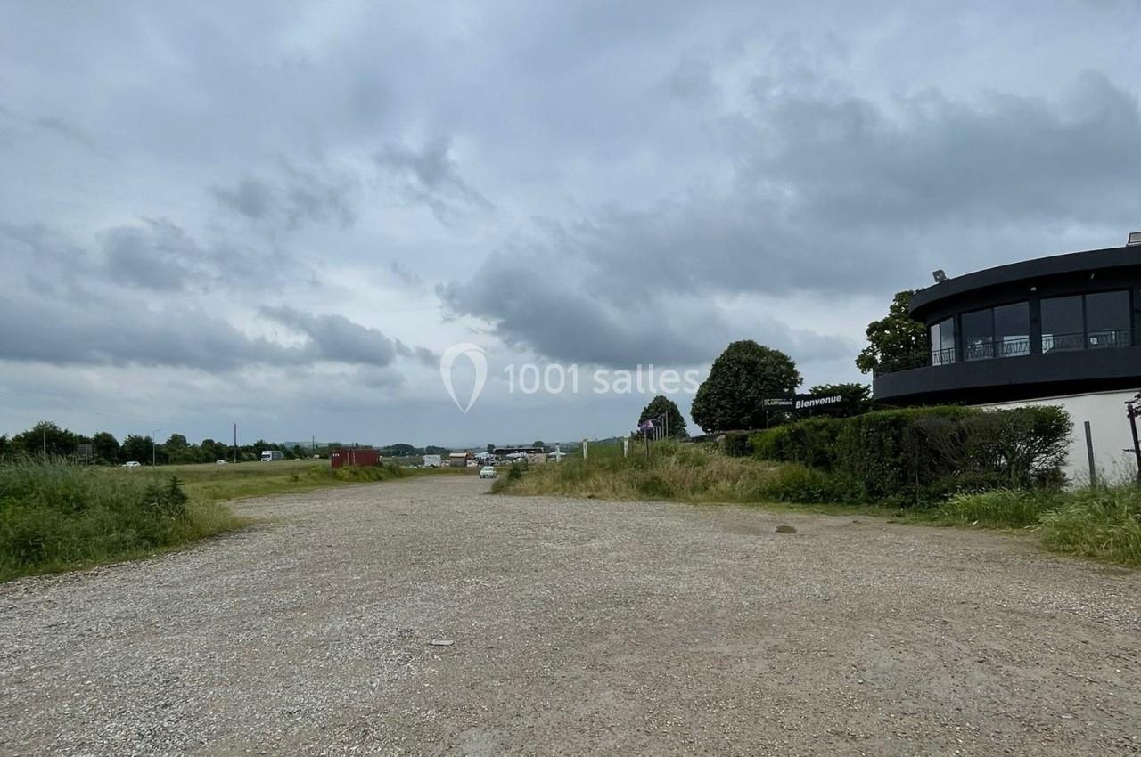 Chemin de gravier bordé de végétation, menant à un bâtiment moderne sous un ciel nuageux.