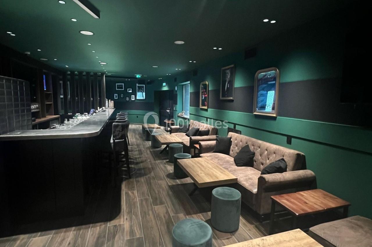 Salle lounge moderne avec canapés, tables basses en bois, tabourets et un bar éclairé dans une ambiance tamisée.