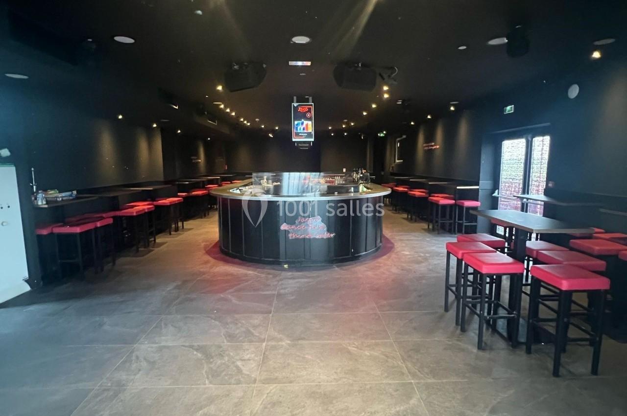 Salle sombre avec un bar central arrondi, entouré de tables hautes et de tabourets rouges.