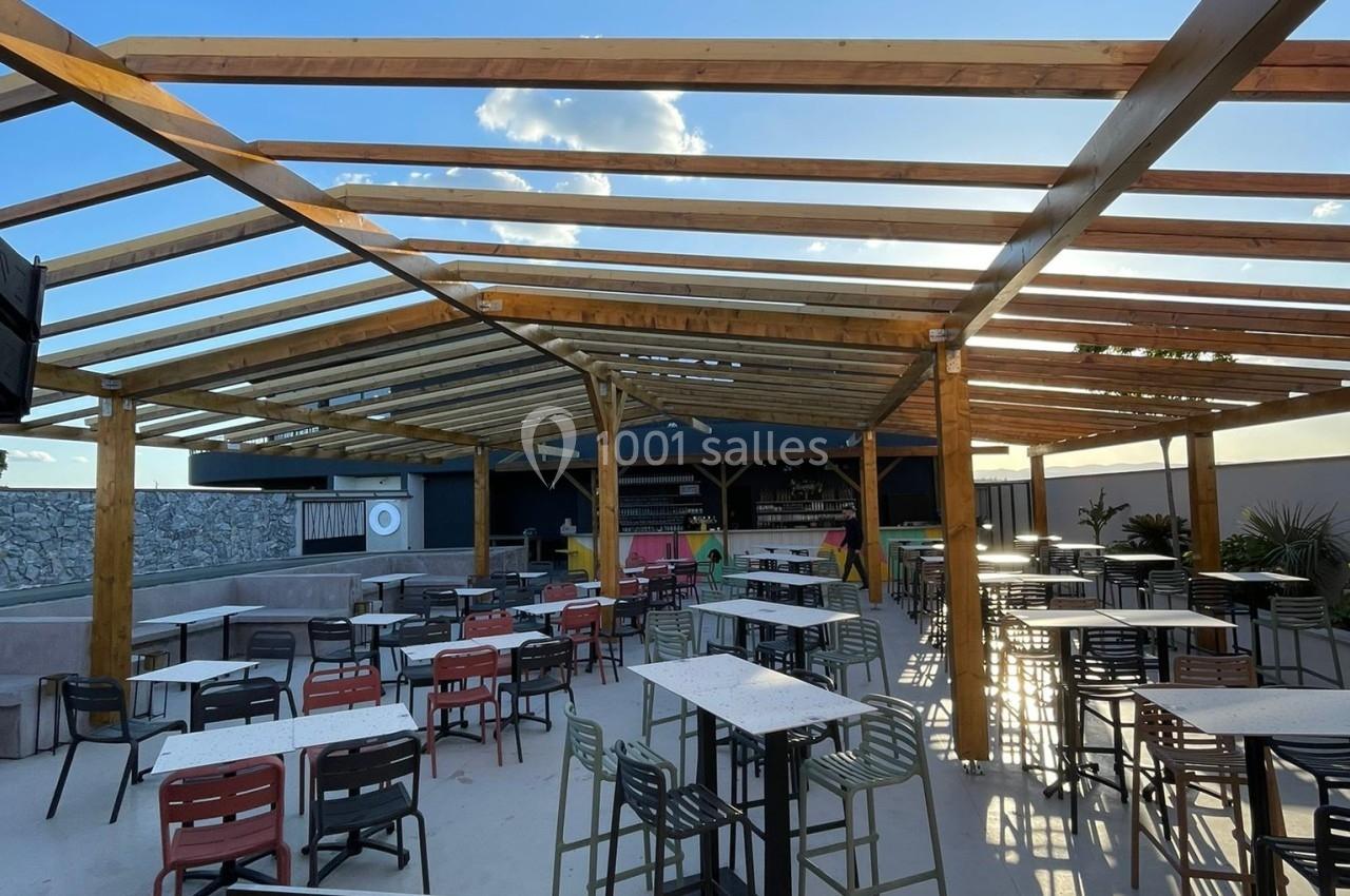 Terrasse extérieure ensoleillée avec tables, chaises et une cabine de DJ au premier plan sous une structure en bois.