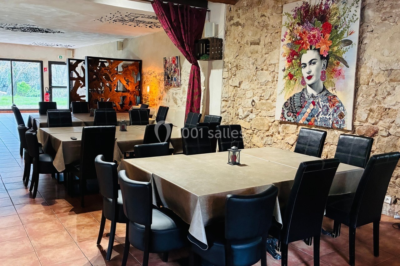 Salle de restaurant avec murs en pierre, tables dressées, chaises noires et tableaux colorés en décoration.