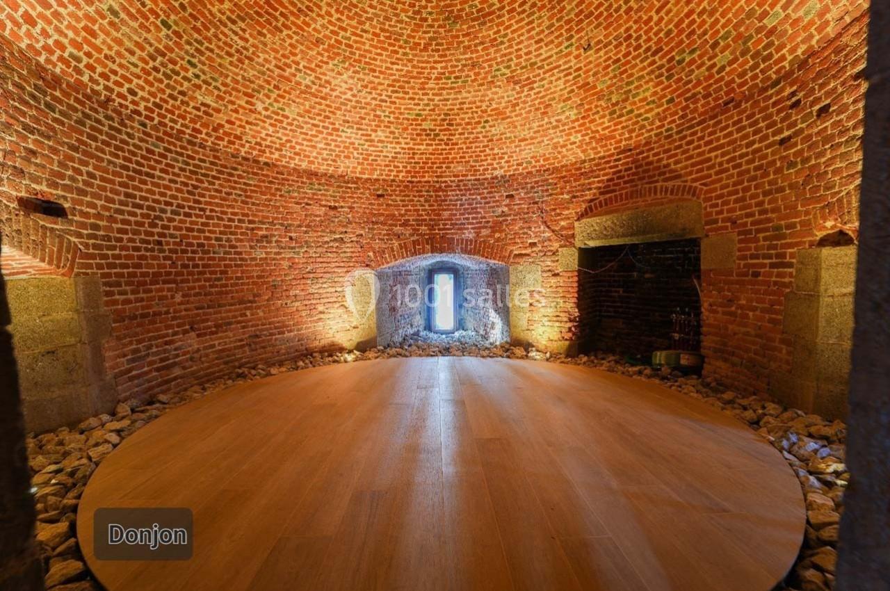 Salle circulaire en briques rouges avec un sol en bois, éclairée par une fenêtre centrale et des lumières murales.