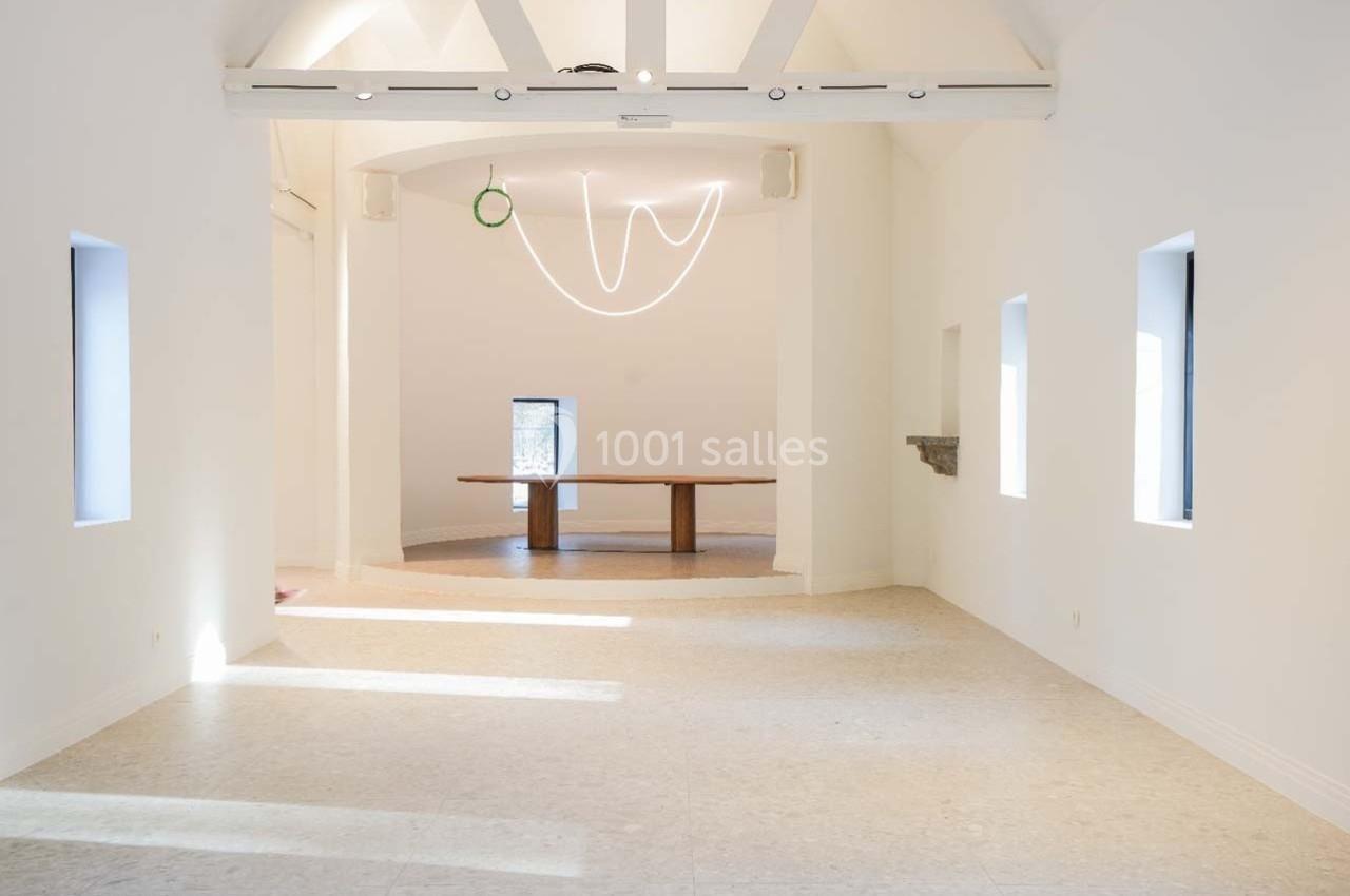Salle lumineuse avec murs blancs, plafond à poutres apparentes, fenêtres étroites et table en bois au fond.
