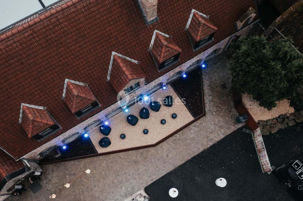 Vue aérienne d'une cour pavée avec des toits en tuiles, des tables rondes et un éclairage bleu disposé en ligne.