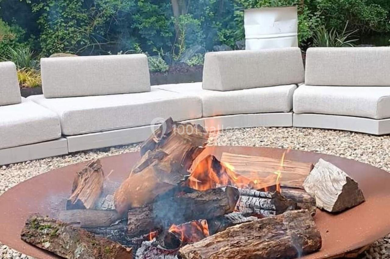 Brasero en métal avec des bûches enflammées, entouré de canapés d'extérieur sur un sol en gravier.