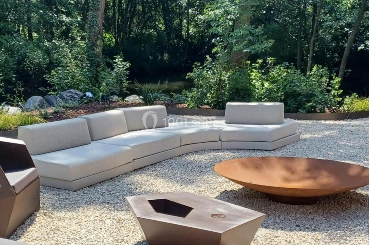 Canapé d'extérieur beige en arc de cercle, posé sur un sol en gravier, avec une grande vasque métallique et une forêt en…