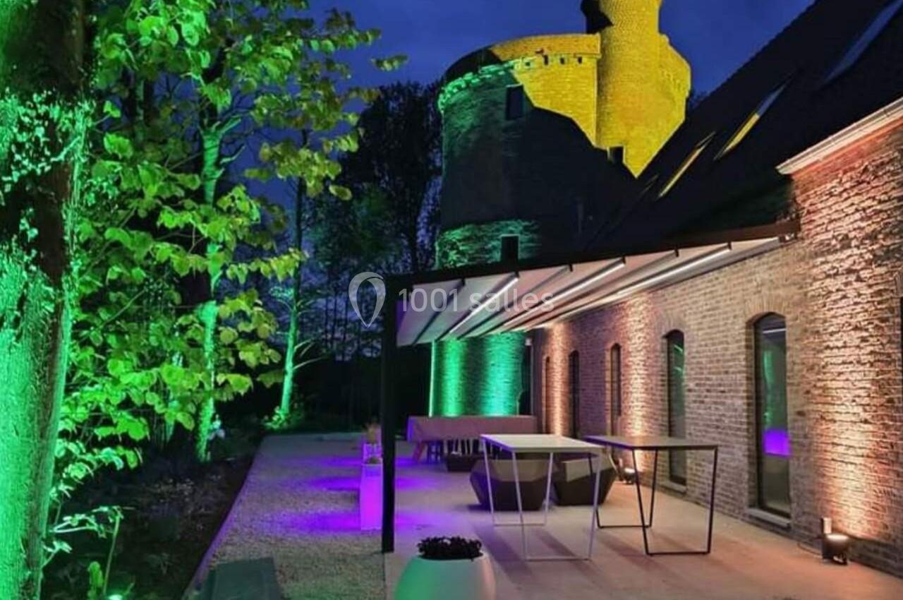 Terrasse éclairée par des lumières colorées devant un bâtiment en pierre, avec une tour illuminée en arrière-plan.