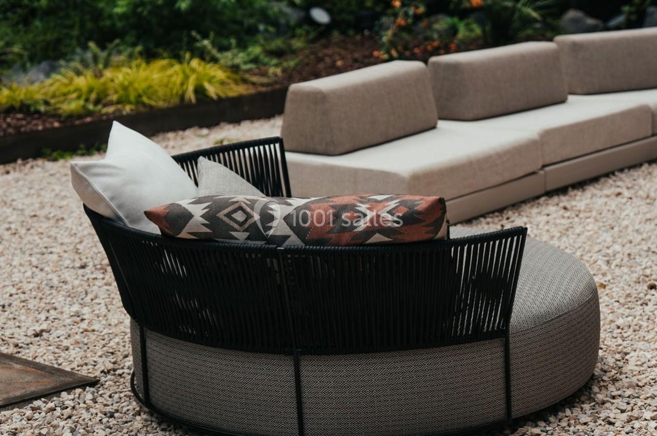 Fauteuil rond en rotin noir avec coussins décoratifs, placé sur un sol en gravier près d'un canapé beige extérieur.