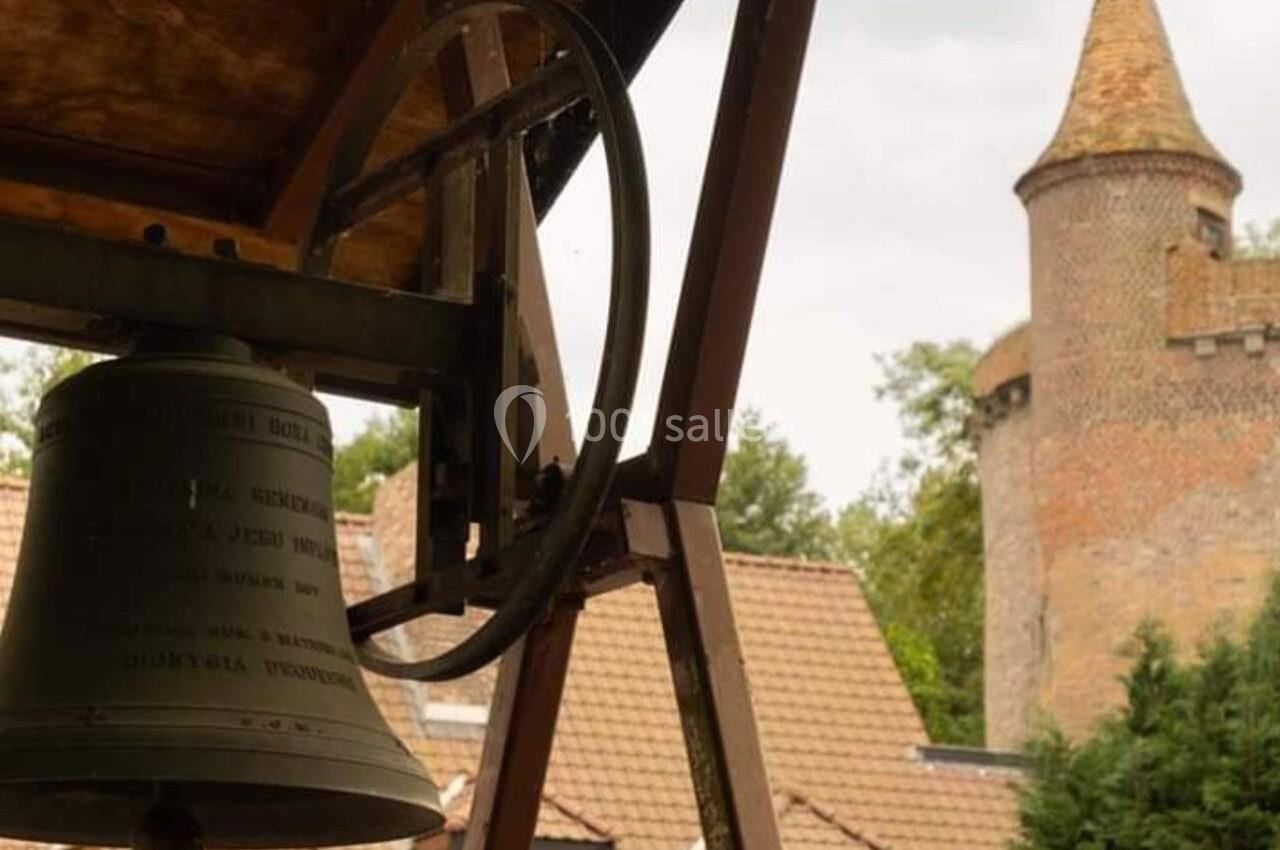 Cloche en métal suspendue sous un abri, avec des toits en tuiles et une tour en briques en arrière-plan.