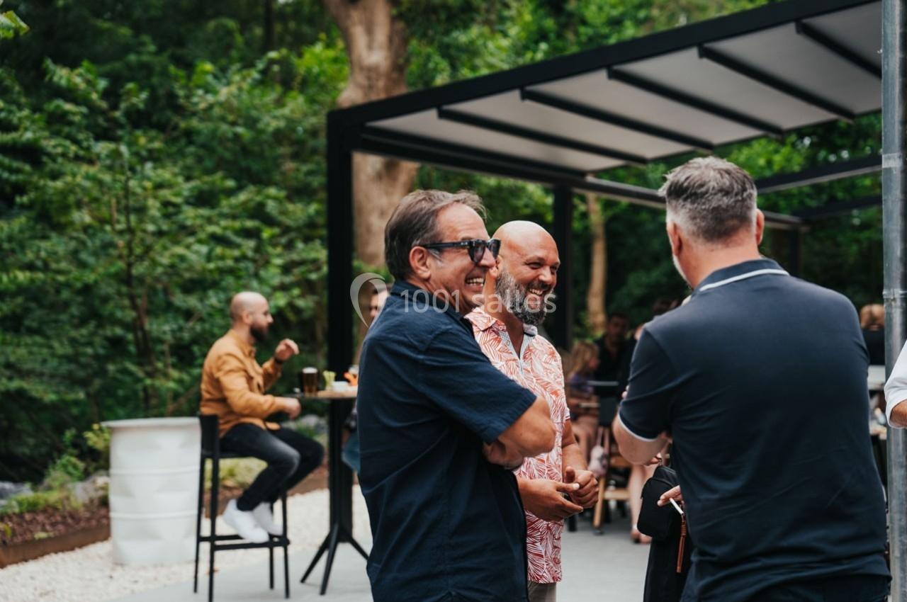 Des personnes discutent debout dans un espace extérieur, entourées de verdure et de mobilier de jardin.
