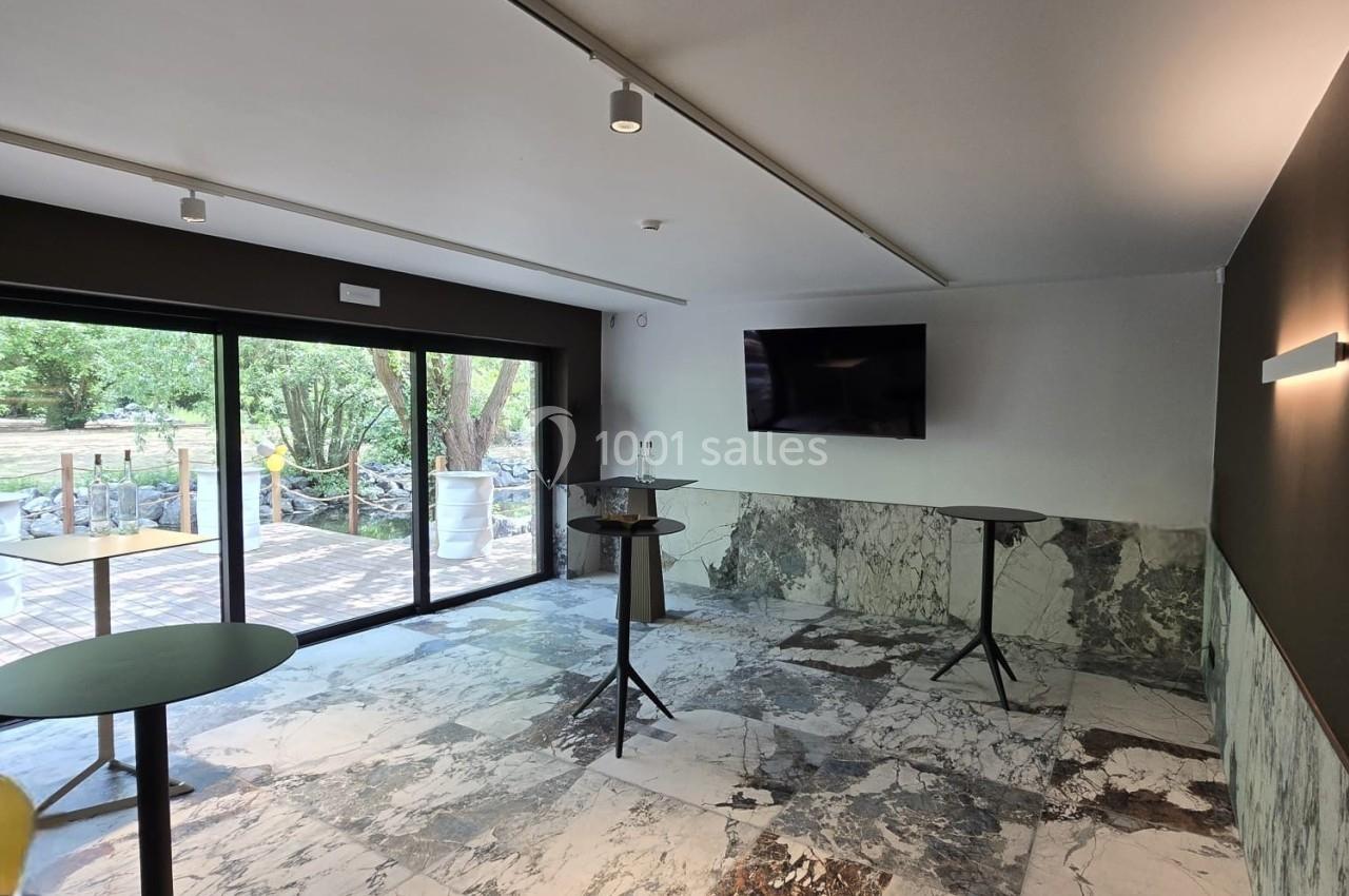 Salle lumineuse avec sol en marbre, tables hautes, écran mural et grandes baies vitrées donnant sur un espace extérieur…