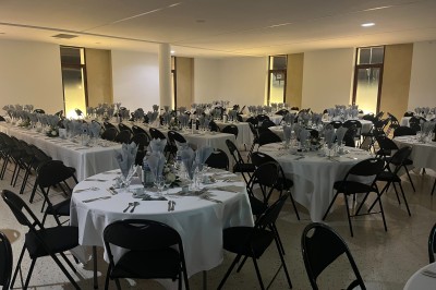 Salle de réception avec tables rondes dressées, nappes blanches, serviettes pliées et chaises noires.