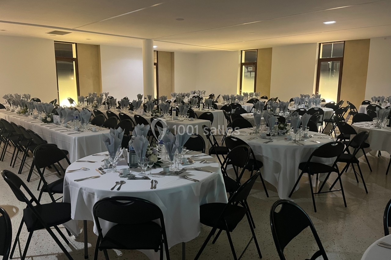 Salle de réception avec des tables rondes dressées, nappes blanches, serviettes pliées et chaises noires.