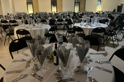 Salle de réception avec tables rondes dressées, nappes blanches, serviettes pliées et chaises noires.