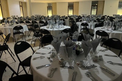 Salle de réception avec tables rondes dressées, nappes blanches, serviettes pliées et chaises noires.