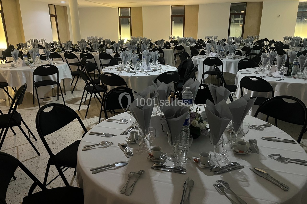 Salle de réception avec tables rondes dressées, nappes blanches, serviettes pliées et chaises noires.