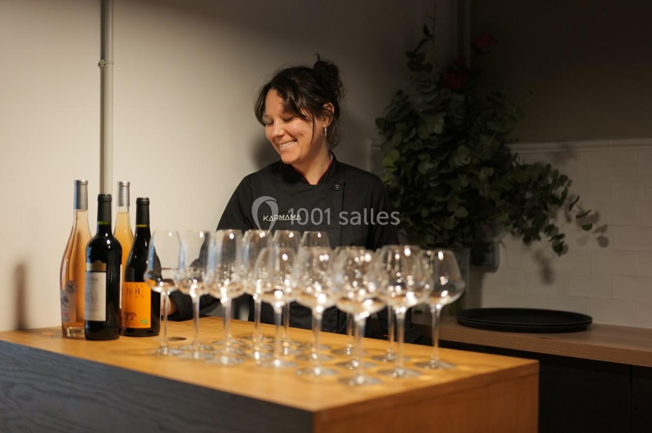 Une femme souriante en tenue professionnelle prépare des verres à vin alignés sur un comptoir en bois.