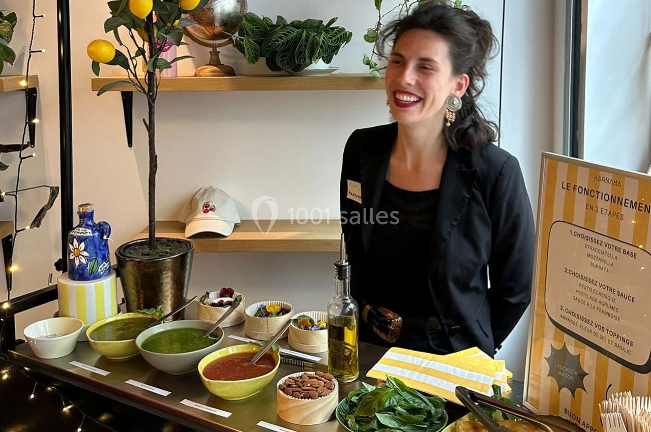 Une femme souriante se tient derrière un stand avec des sauces, des ingrédients frais et un panneau explicatif.