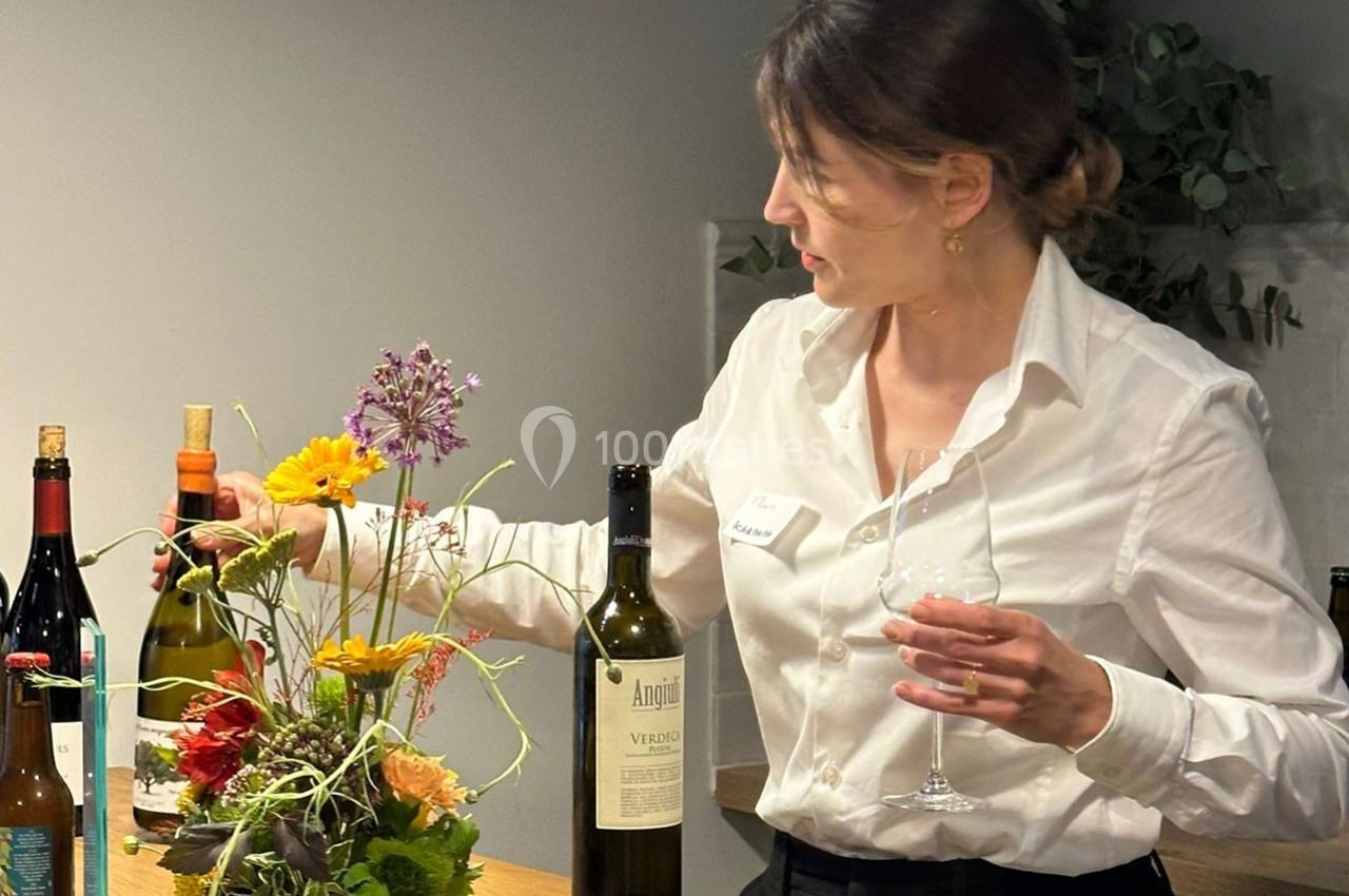 Une femme en chemise blanche sert du vin près d'un bouquet de fleurs colorées sur un comptoir en bois.
