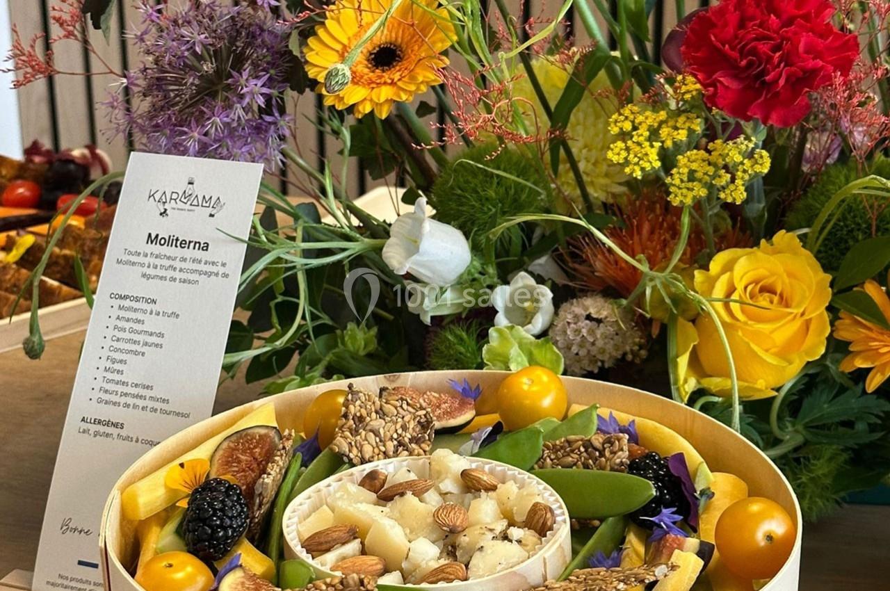 Plateau de fromage garni de fruits secs, fleurs comestibles et légumes, entouré d'un décor floral coloré.