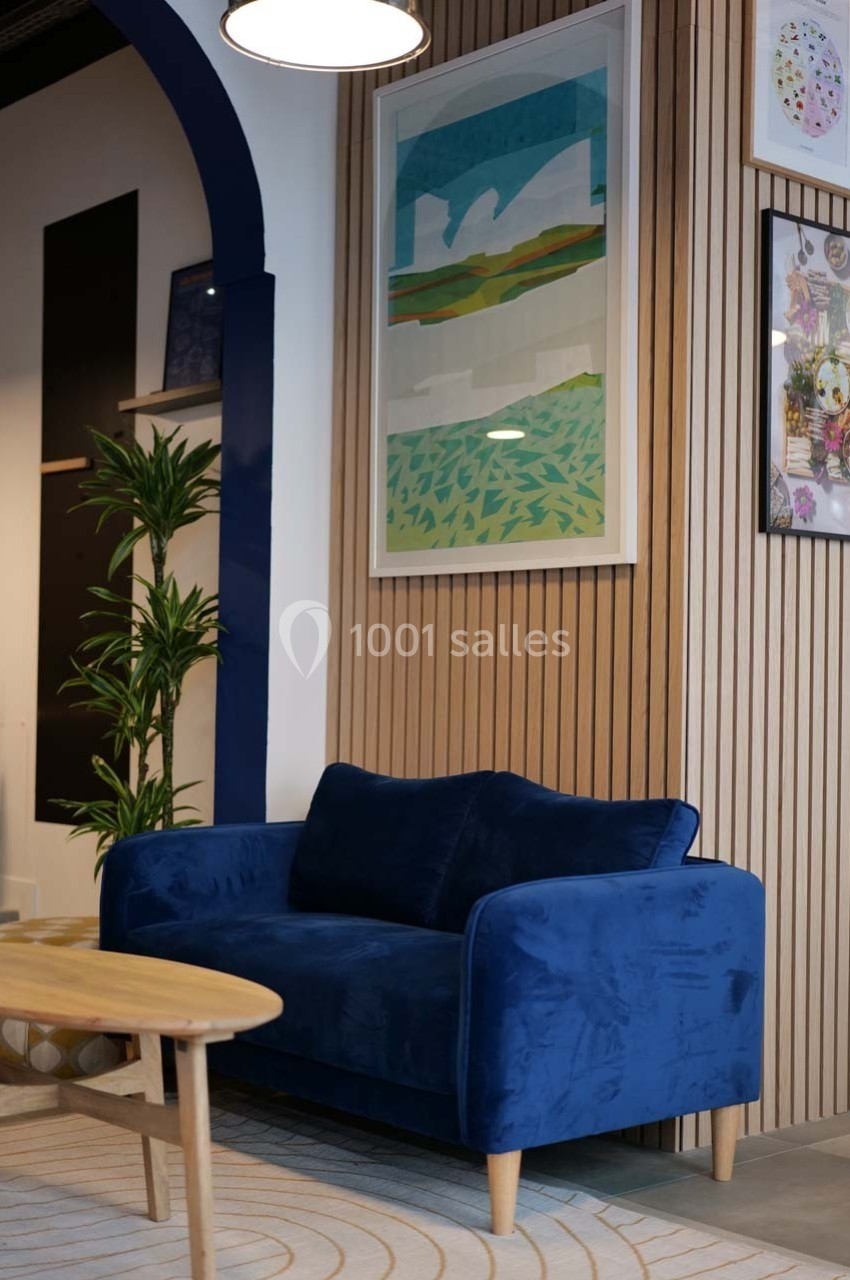 Canapé bleu en velours devant un mur en bois avec des cadres décoratifs, éclairé par une suspension.