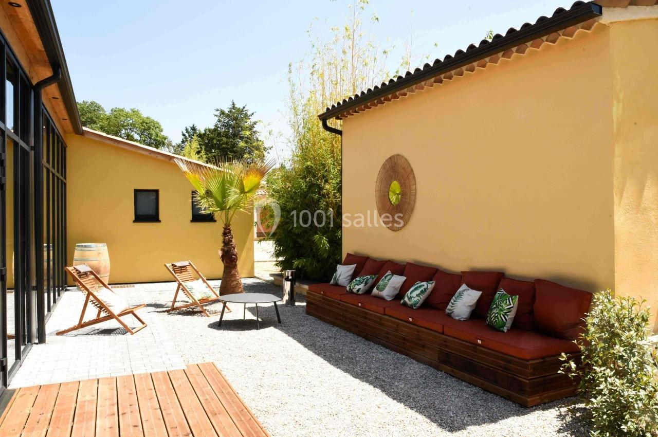 Terrasse ensoleillée avec banc en bois, coussins colorés, chaises longues, palmiers et murs jaunes.