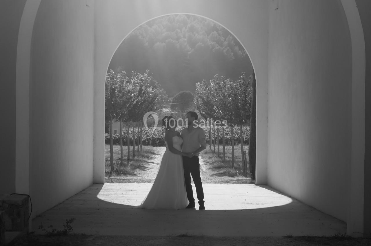 Un couple debout sous une arche, éclairé par un rayon de lumière, avec des arbres alignés en arrière-plan.