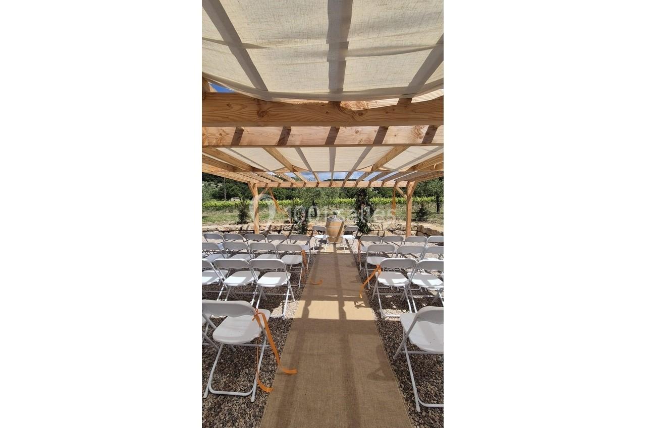 Chaises blanches alignées sous une pergola en bois, disposées pour une cérémonie en extérieur.