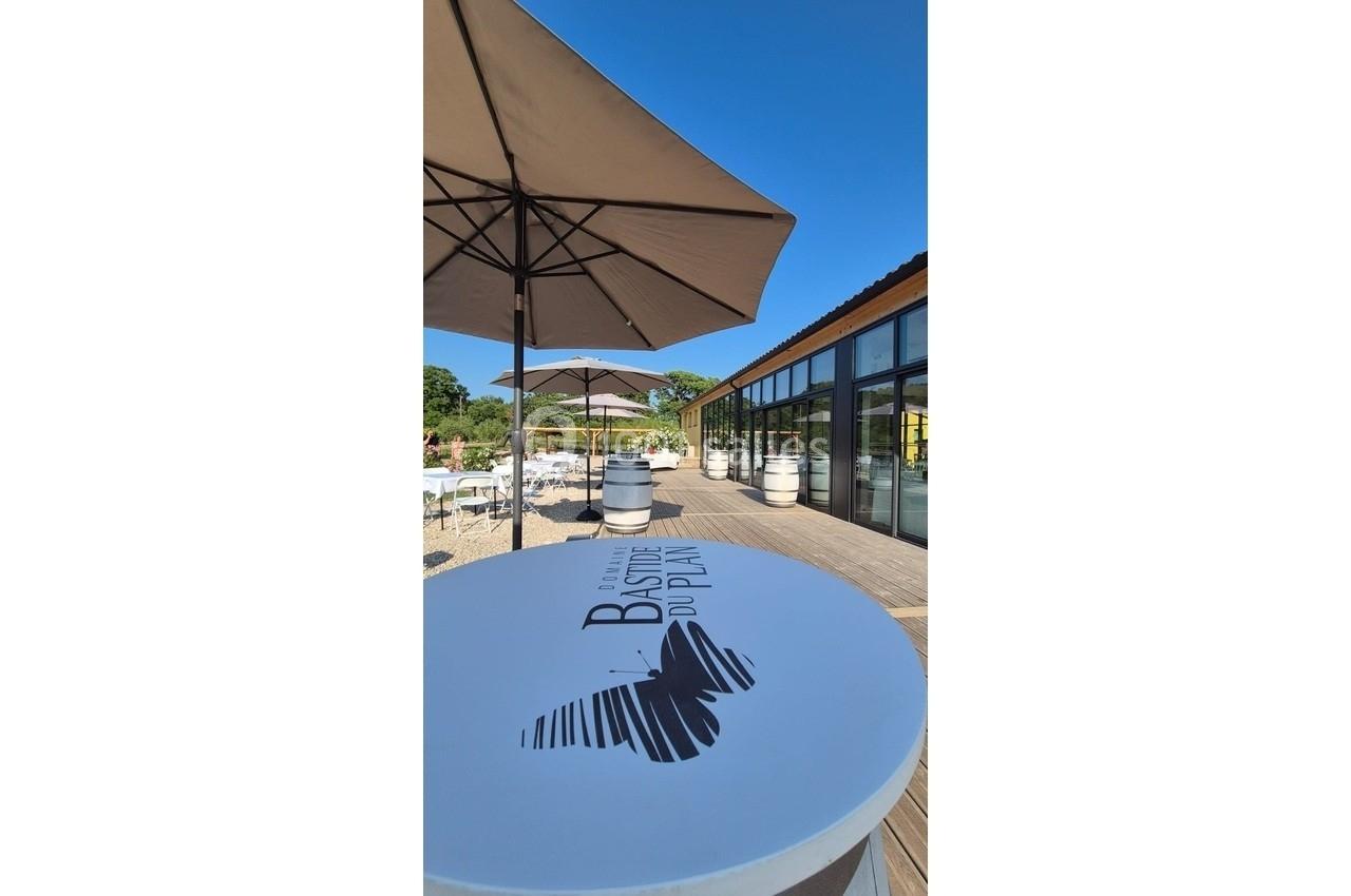 Terrasse ensoleillée avec parasols, tables et barils, devant une façade vitrée moderne entourée de verdure.