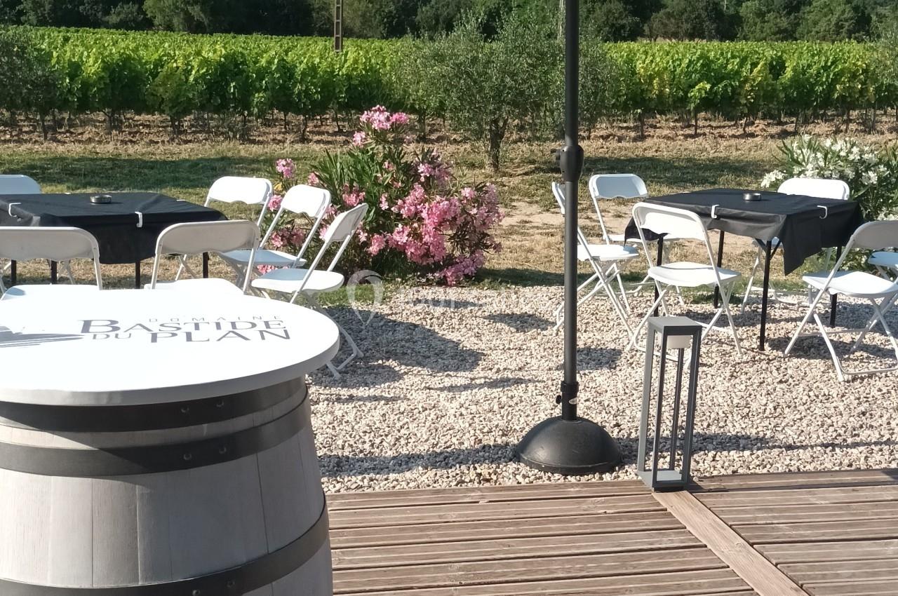 Terrasse en bois avec tonneau en table, parasol, chaises pliantes et vue sur des vignes et collines verdoyantes.