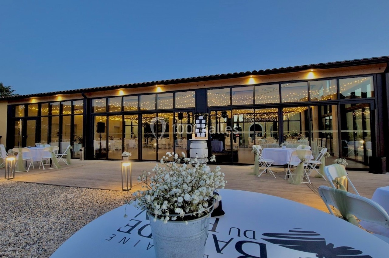 Salle de réception vitrée illuminée en soirée, avec tables décorées en extérieur et guirlandes lumineuses visibles à l…