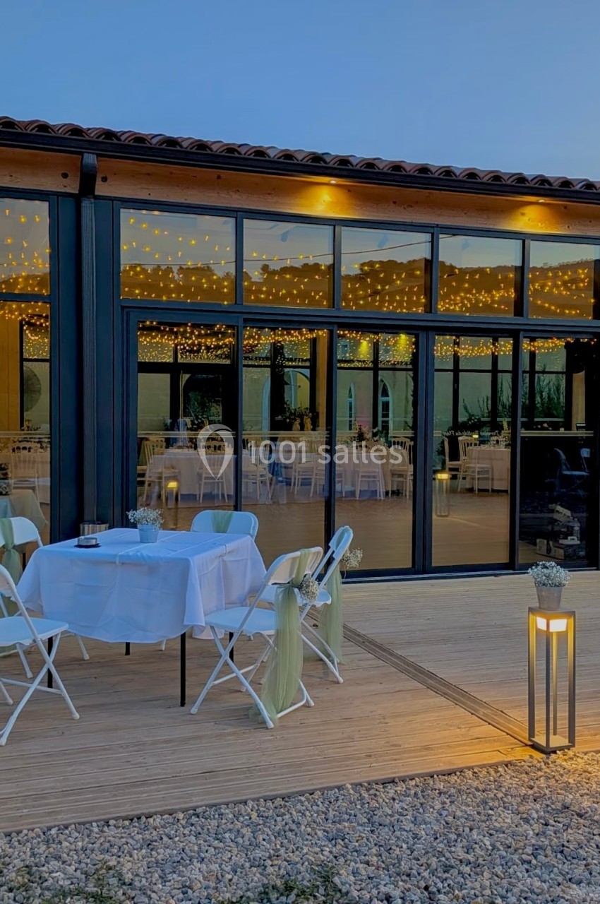 Salle vitrée éclairée par des guirlandes lumineuses, avec une terrasse aménagée de tables et chaises décorées.