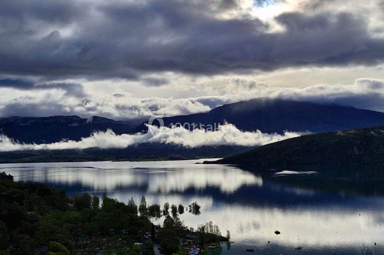 Paysage lacustre entouré de collines, avec des nuages bas et une lumière diffuse reflétée sur l'eau calme.