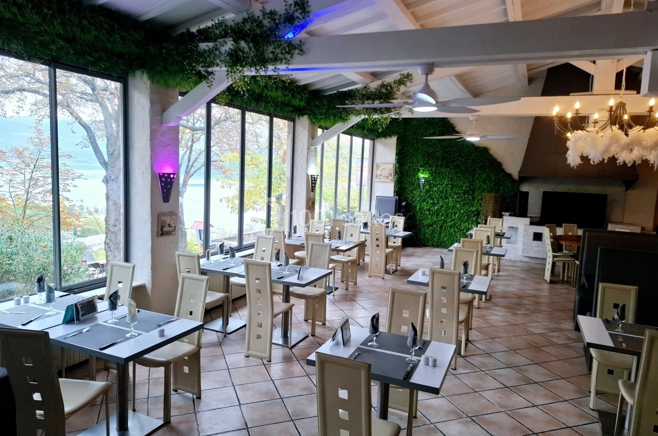 Salle de restaurant lumineuse avec de grandes baies vitrées, des tables dressées et des murs décorés de végétation.