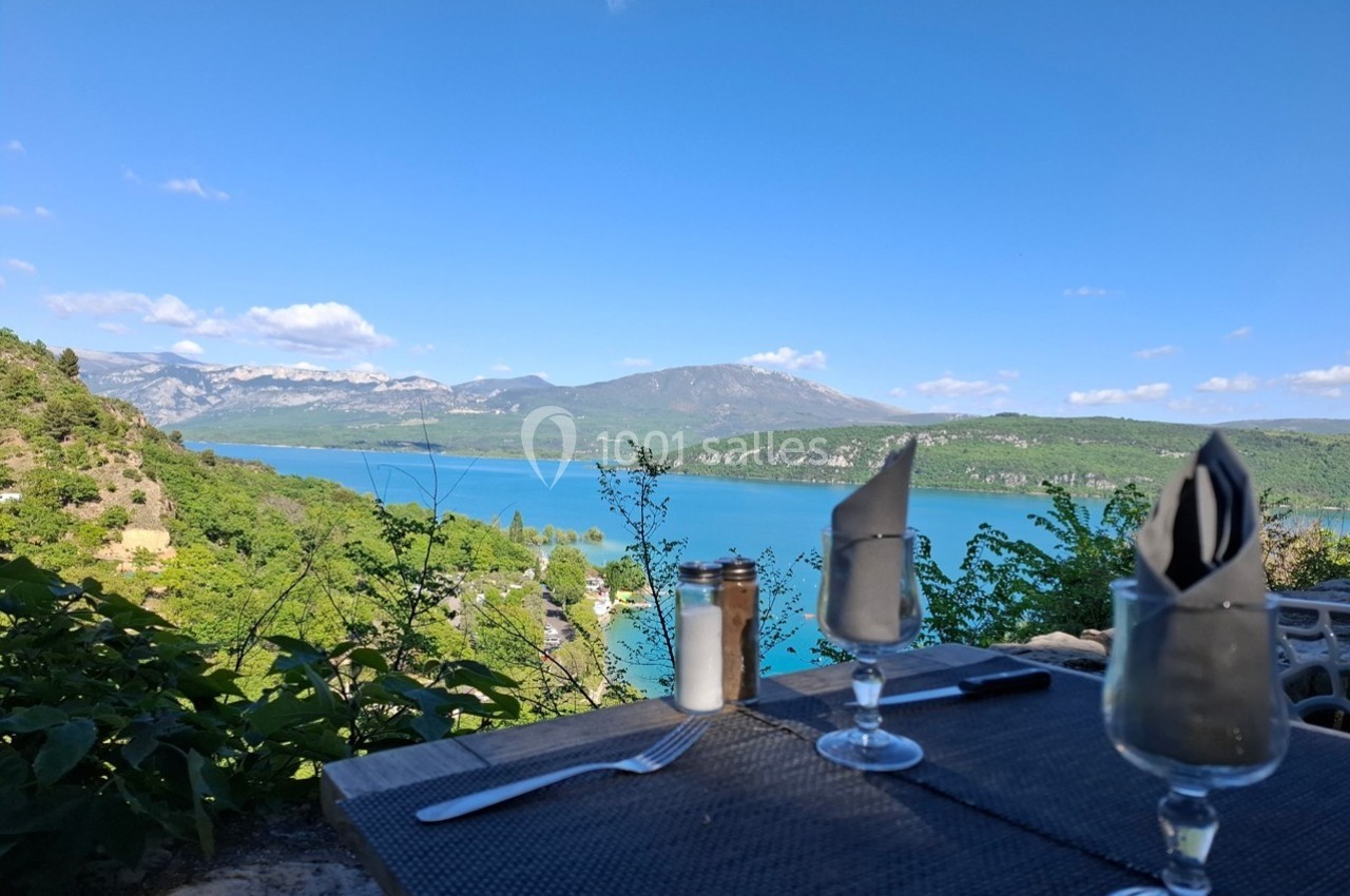 Table dressée avec vue sur un lac turquoise entouré de montagnes et de végétation sous un ciel dégagé.