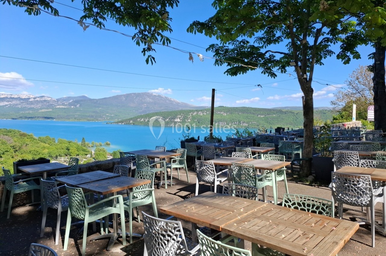 Terrasse de restaurant avec tables et chaises, offrant une vue sur un lac entouré de montagnes sous un ciel bleu.