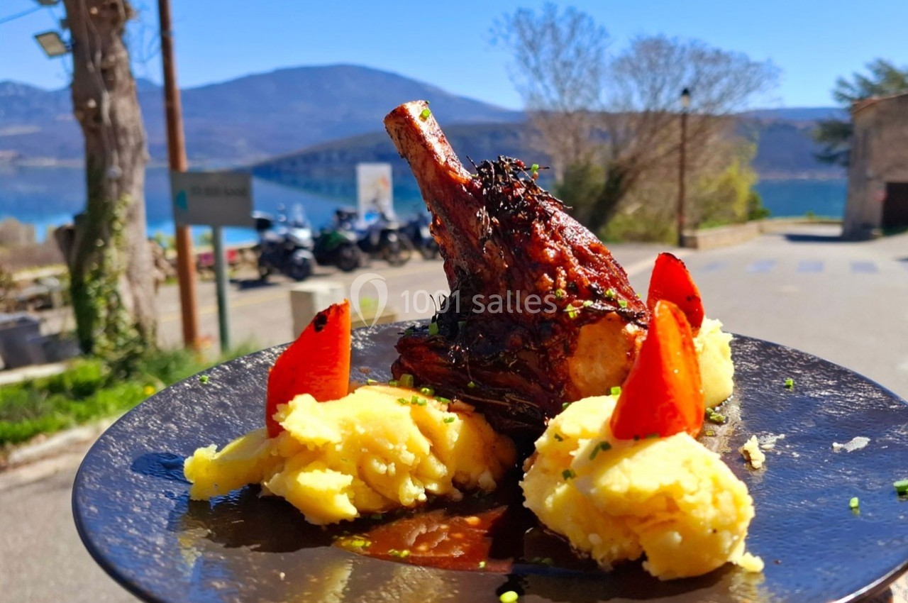 Plat de viande braisée avec purée et légumes, servi en extérieur avec vue sur un lac et des montagnes en arrière-plan.