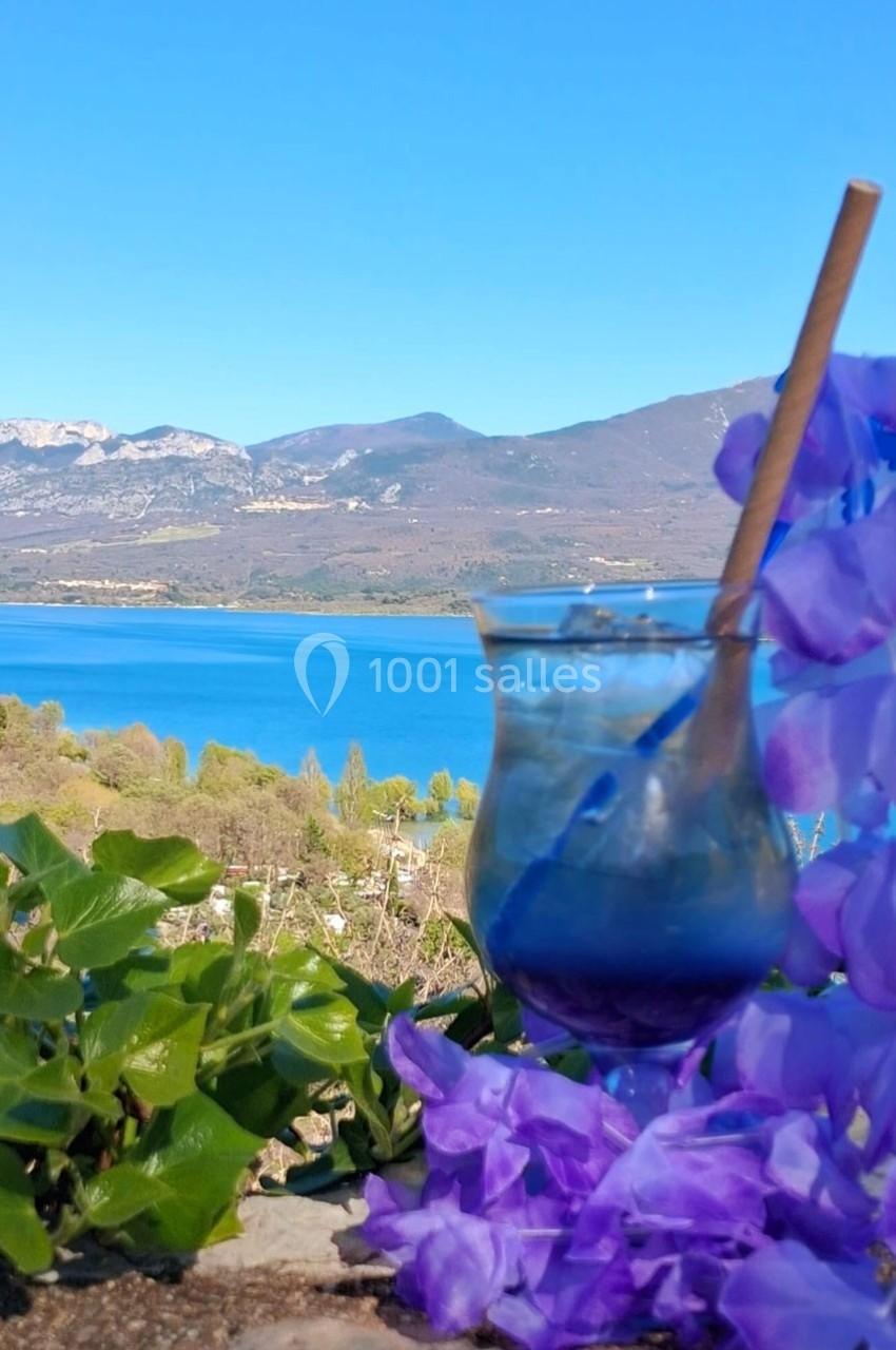 Verre avec paille entouré de fleurs violettes, posé devant un lac et des montagnes sous un ciel bleu.