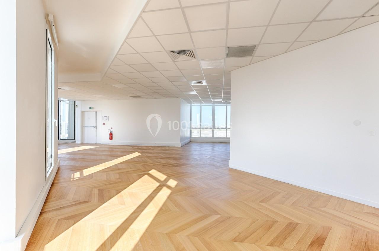 Bureau vide avec parquet en bois clair, murs blancs et plafond à dalles, éclairé par de grandes fenêtres.
